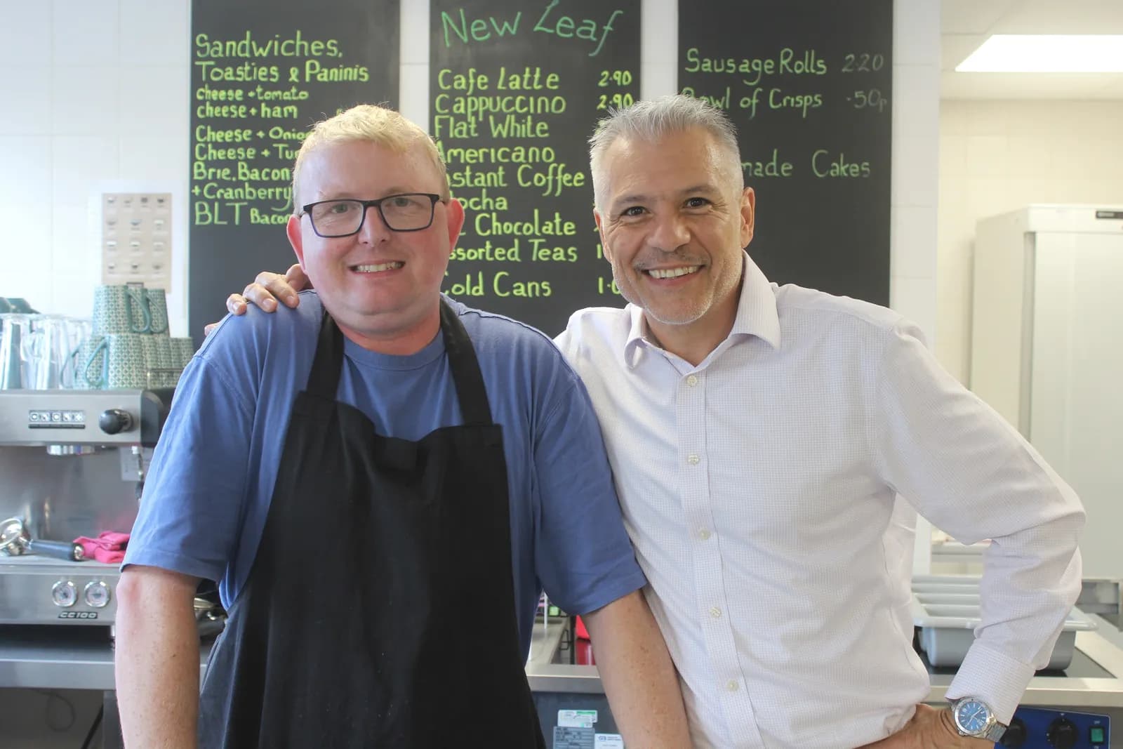 Two smiling men, one in a blue shirt and black apron and the other in a white button-up shirt, stand in what appears to be a cafe in front of chalkboards listing menu items.