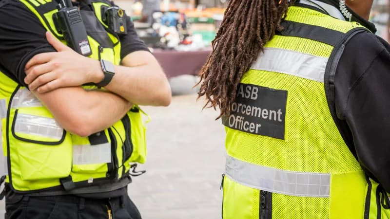 Two people in high-visibility vests are standing next to each other, one with crossed arms and a radio clipped to their vest, and the other with dreadlocks and a patch on their back reading "ASB Enforcement Officer."