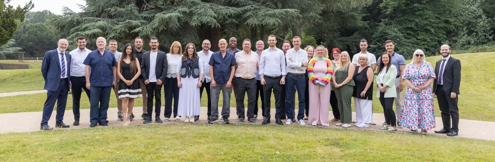 A diverse group of men and women, varying in age and ethnicity, stand together on a grassy lawn with trees in the background, many smiling at the camera.