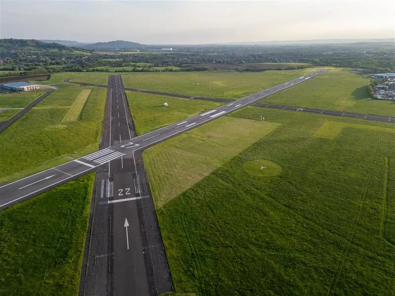 An aerial view of an airport with intersecting black runways marked with white lines, surrounded by green grass fields and distant hills.