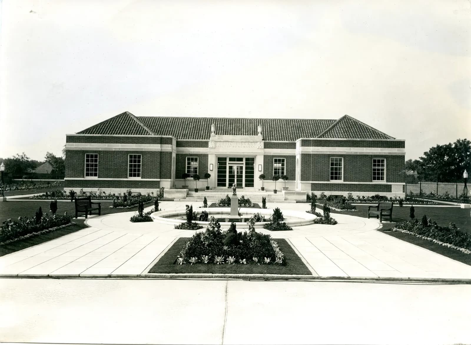 A grand, symmetrical brick building with a tiled roof, featuring a central entrance with a fountain and manicured gardens in the foreground.