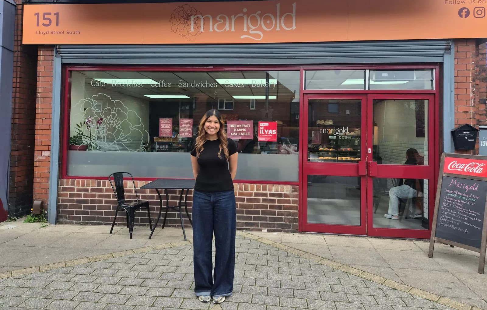 A woman stands smiling directly in front of the Marigold cafe entrance, which has a large window displaying cakes and red-framed double doors. A small table and two chairs are visible outside the cafe entrance.