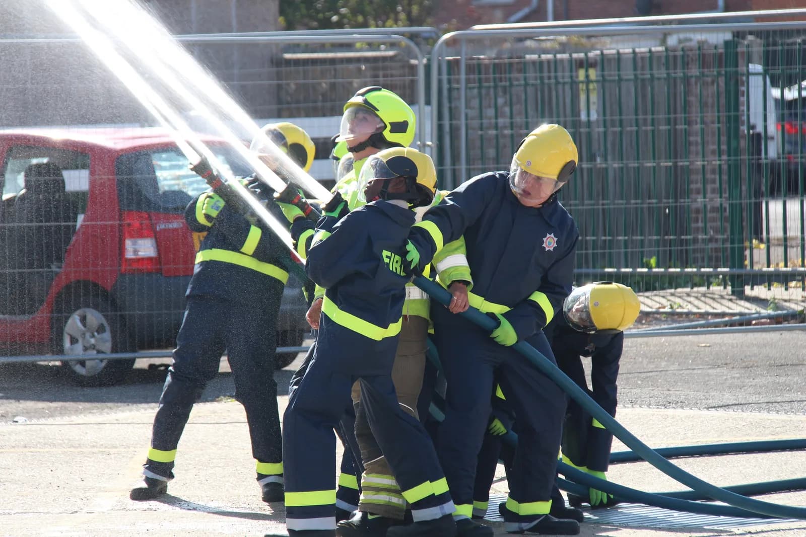 A group of children wearing firefighter suits and yellow helmets are holding a hose and spraying water. A red car is visible in the background behind a metal fence.