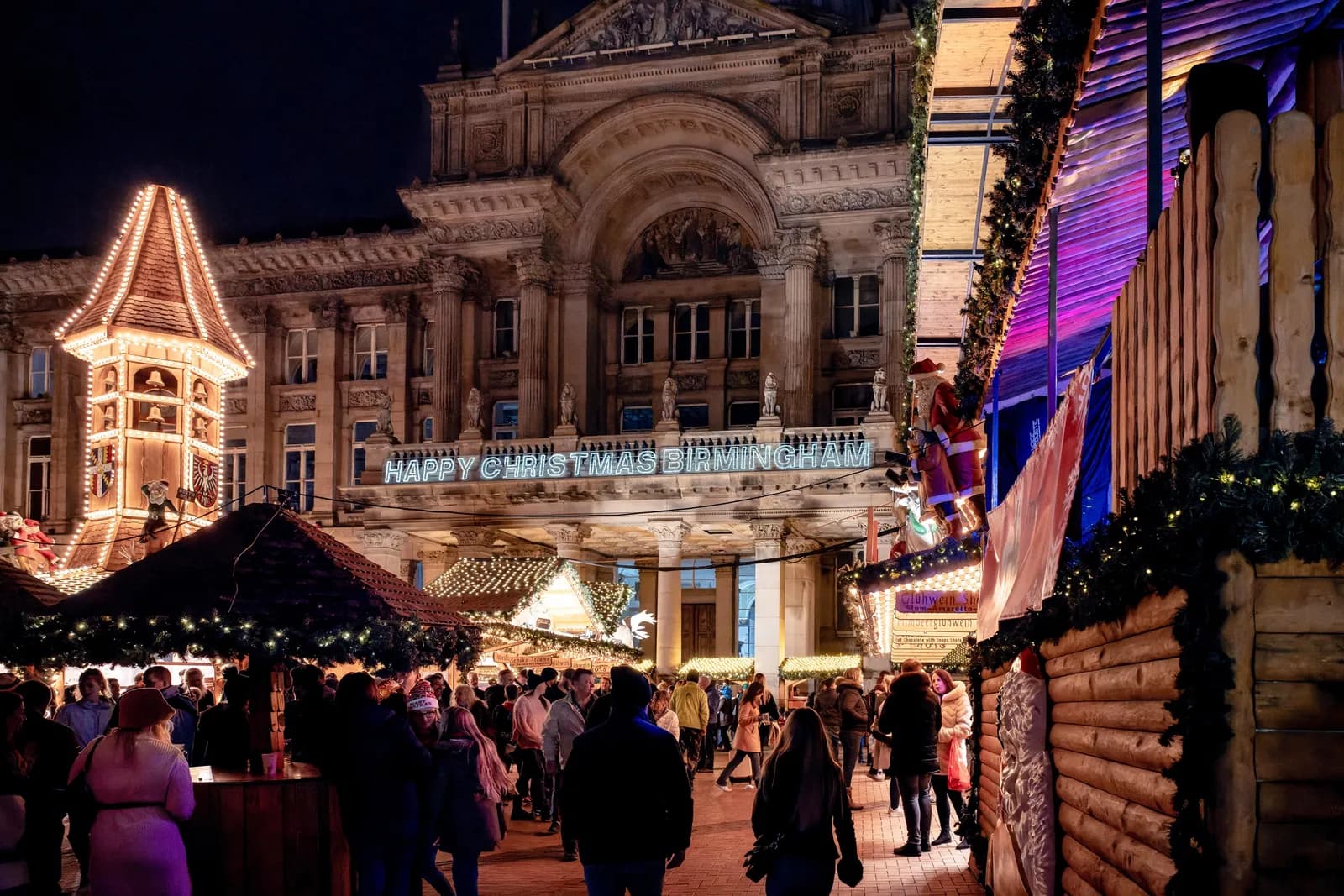 A bustling Christmas market in Birmingham at night, with a large ornate building in the background featuring a lit-up sign that reads "HAPPY CHRISTMAS BIRMINGHAM".