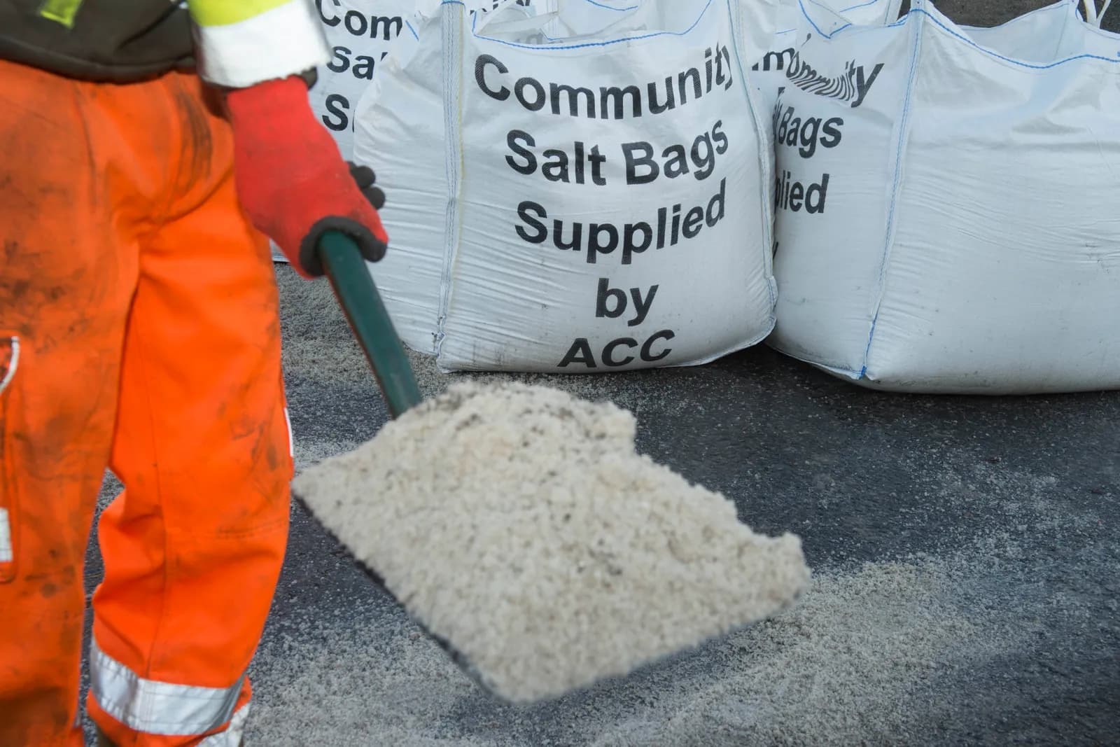 A person wearing orange trousers and red gloves holds a shovel full of salt next to several large white bags of salt labeled "Community Salt Bags Supplied by ACC."
