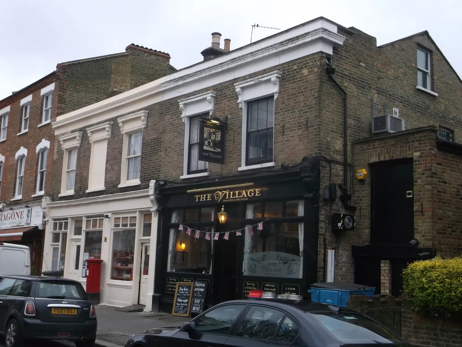 A red brick building housing "The Village" pub is seen from the street, with parked cars in the foreground.