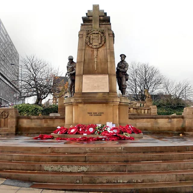 A stone war memorial with two soldier statues, topped by a cross, and surrounded by steps and poppy wreaths.
