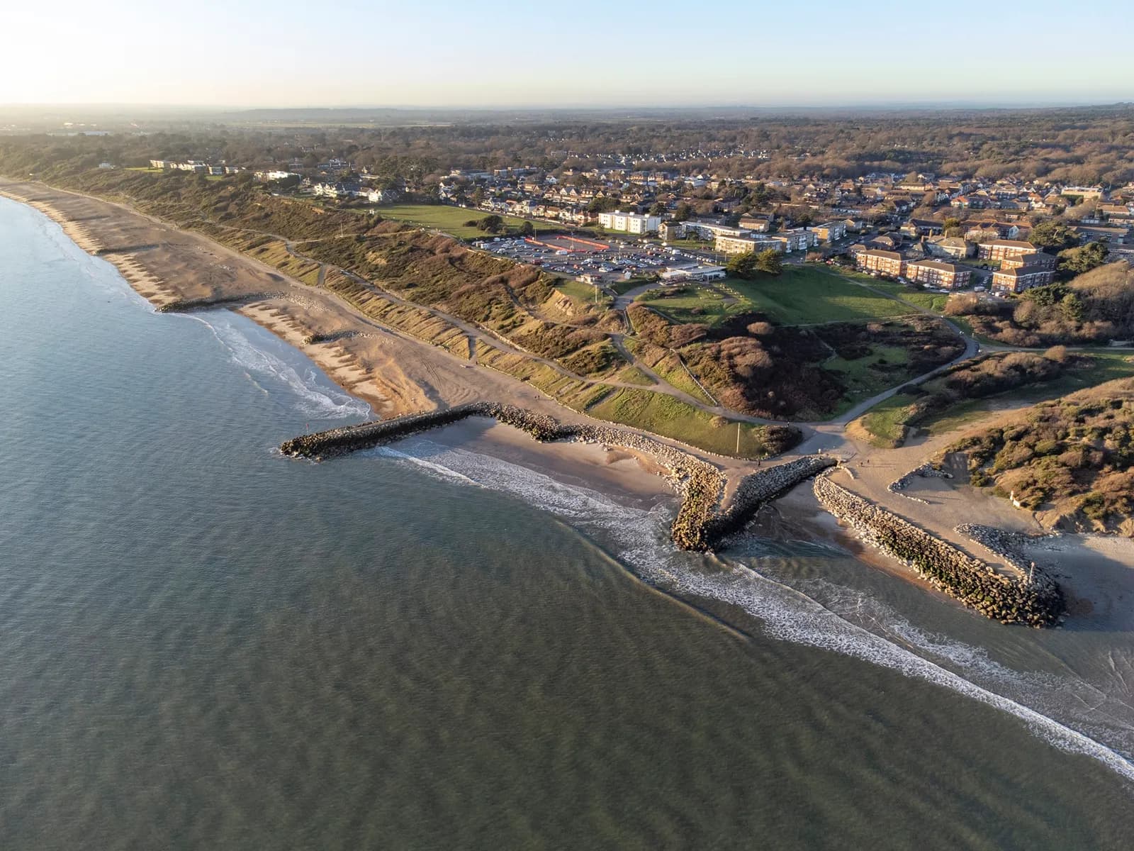 An aerial view captures the curving coastline where a sandy beach meets the ocean, with several rock groynes extending into the water and a residential area visible on the green hills above the shore.