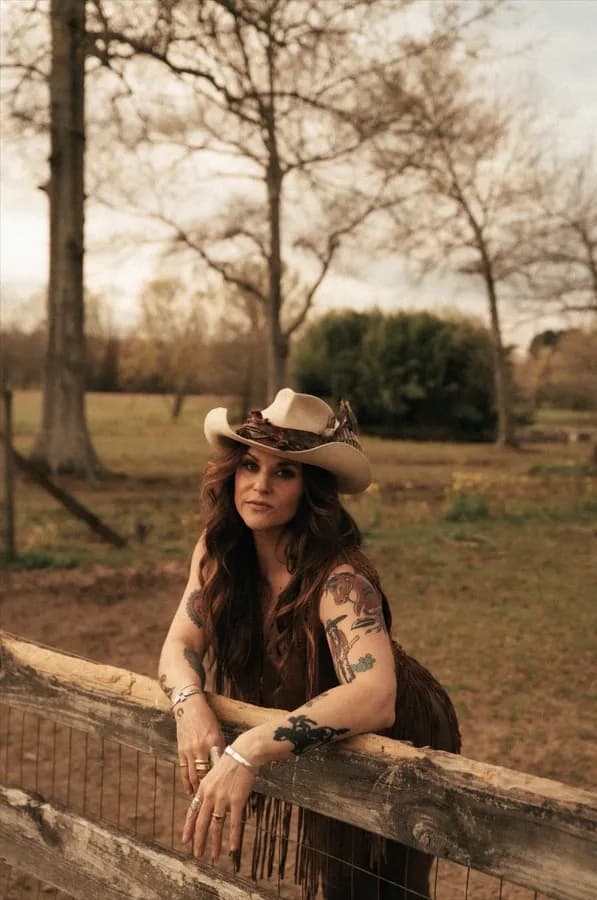 A woman wearing a cowboy hat and a fringed top, leaning on a wooden fence with trees and a bush in the background, showcasing her arm tattoos.