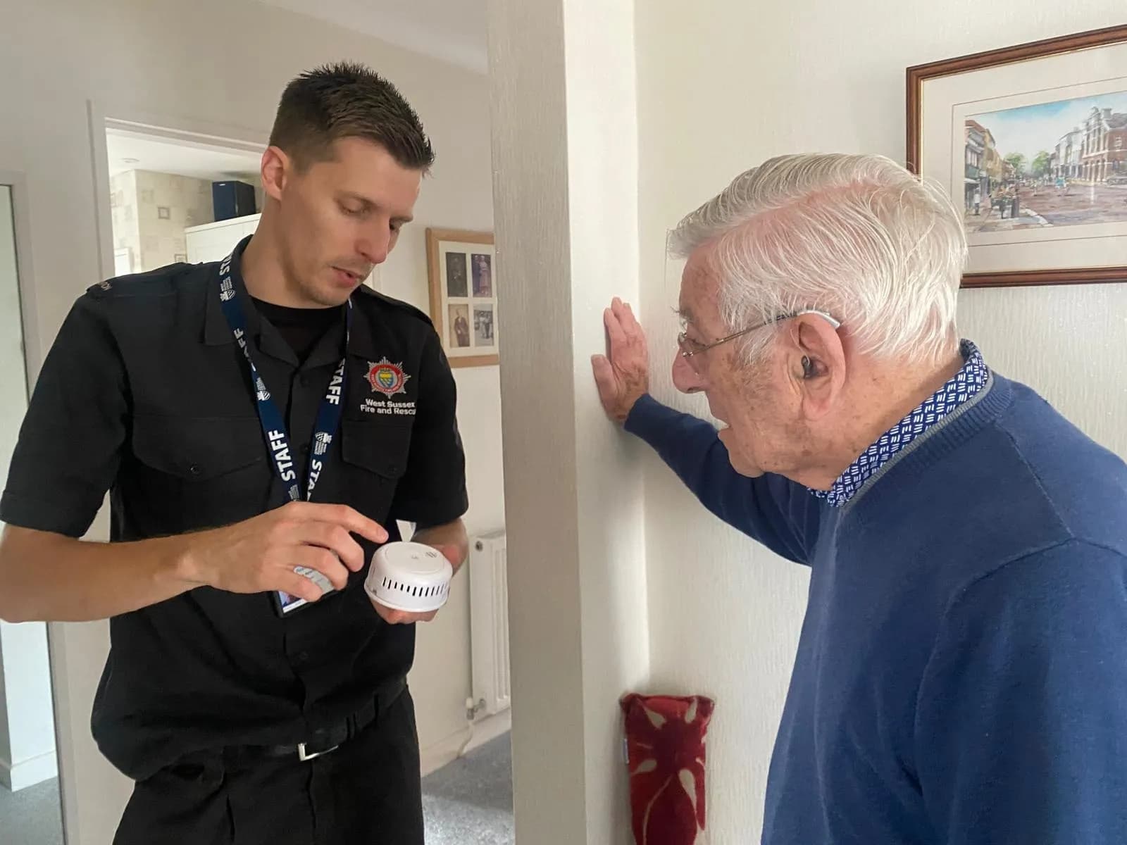 A fire department staff member in a black uniform shows a white smoke detector to an elderly man with glasses and a hearing aid who is leaning against a white wall.