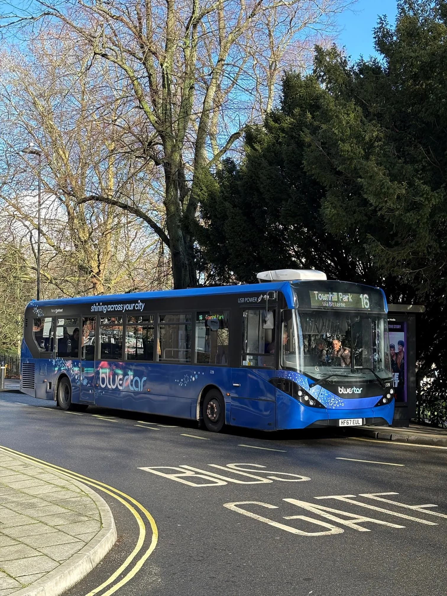 A bright blue bus with 'bluestar' branding and the number '16' on its display is parked on a street with a 'BUS GATE' marking, surrounded by trees.