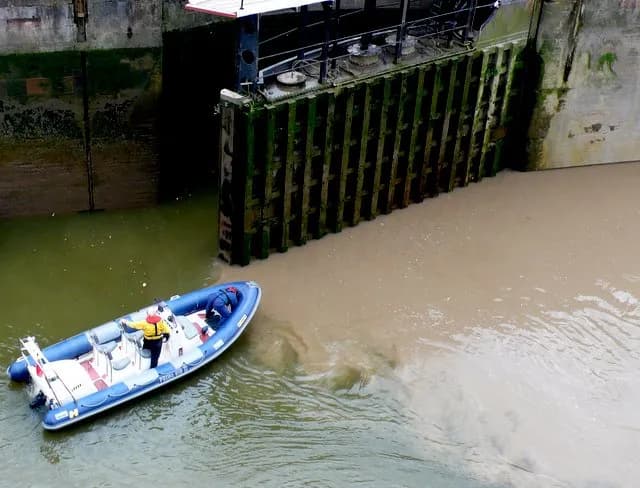 A blue and white inflatable boat with two people on board navigates muddy water near a moss-covered lock gate.