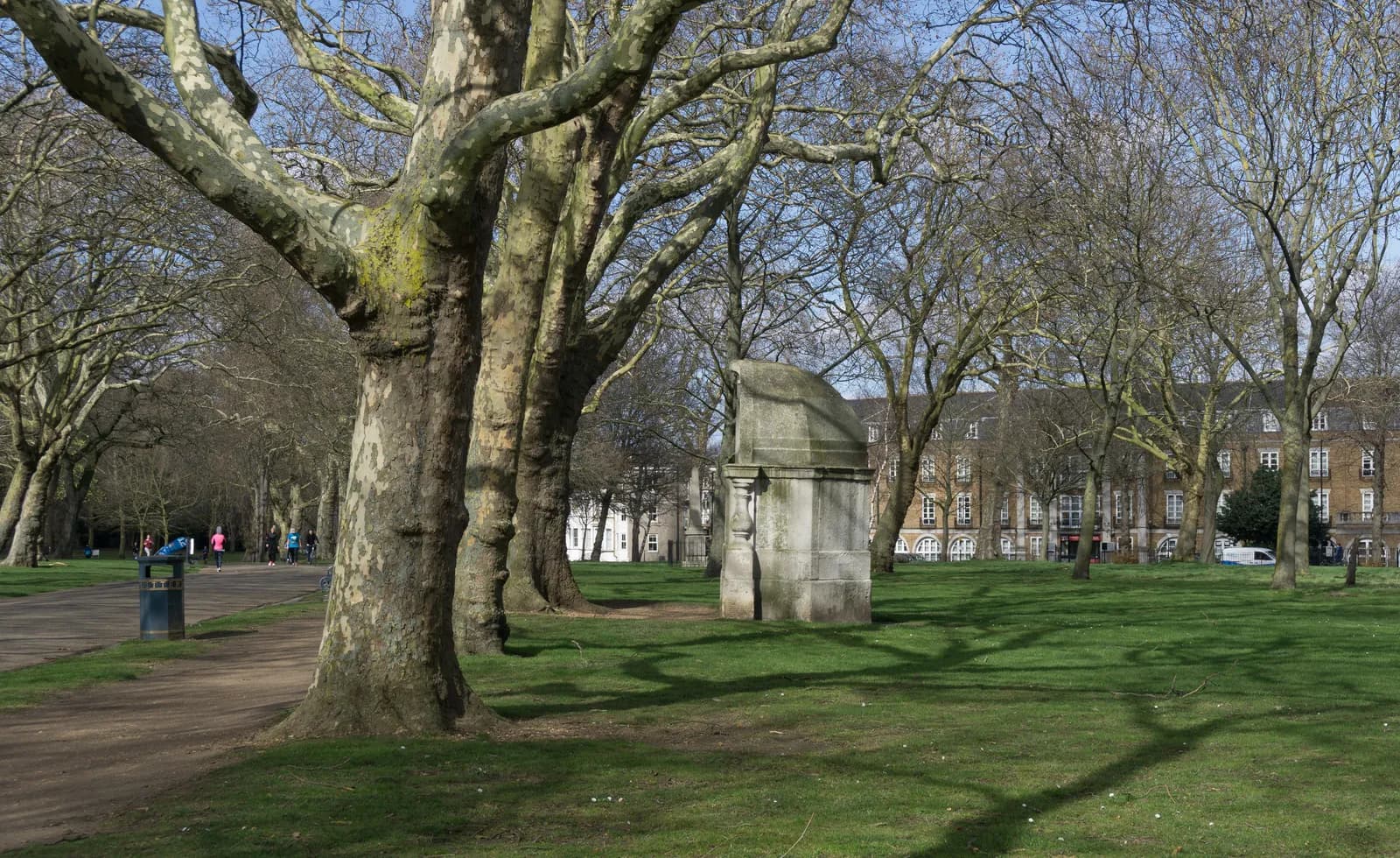 A park scene with large, bare trees forming an avenue, leading to distant buildings, with a stone monument and a few people in the background.