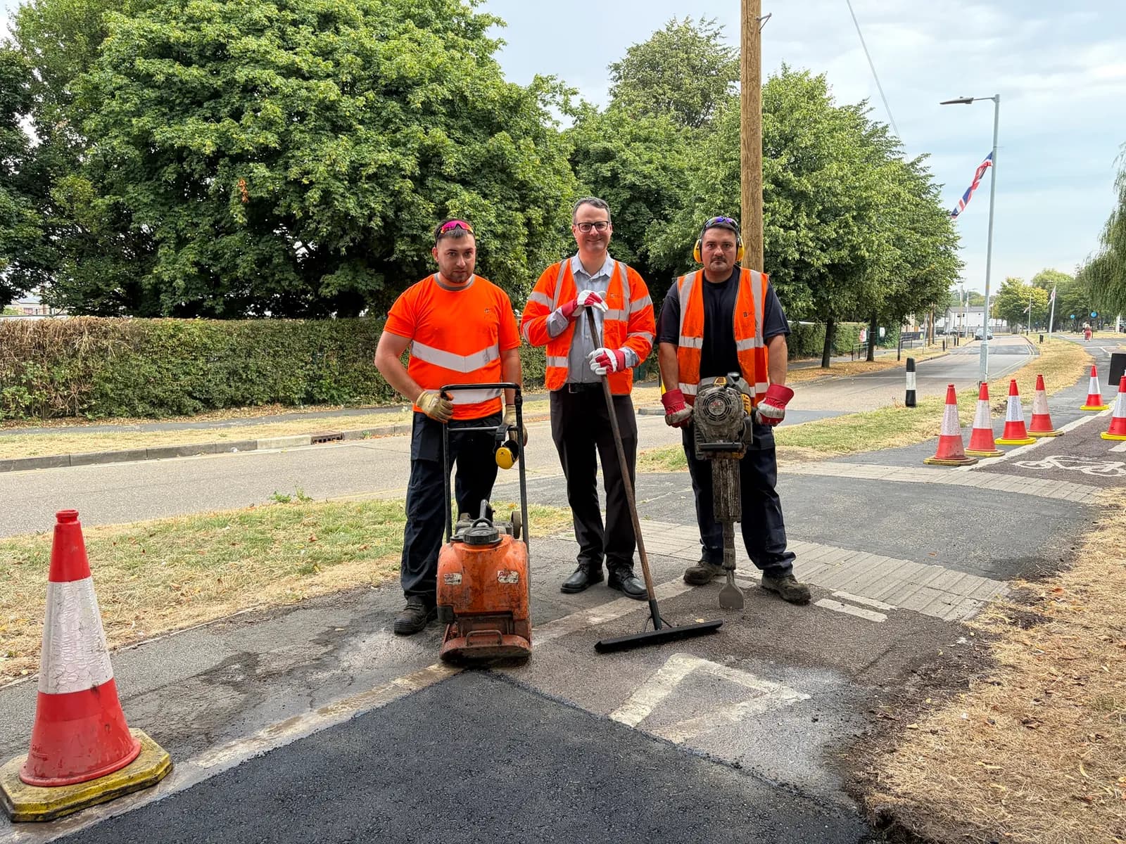 Three men in high-visibility clothing stand on a freshly paved patch of asphalt next to construction equipment and orange cones, with trees and a road in the background.