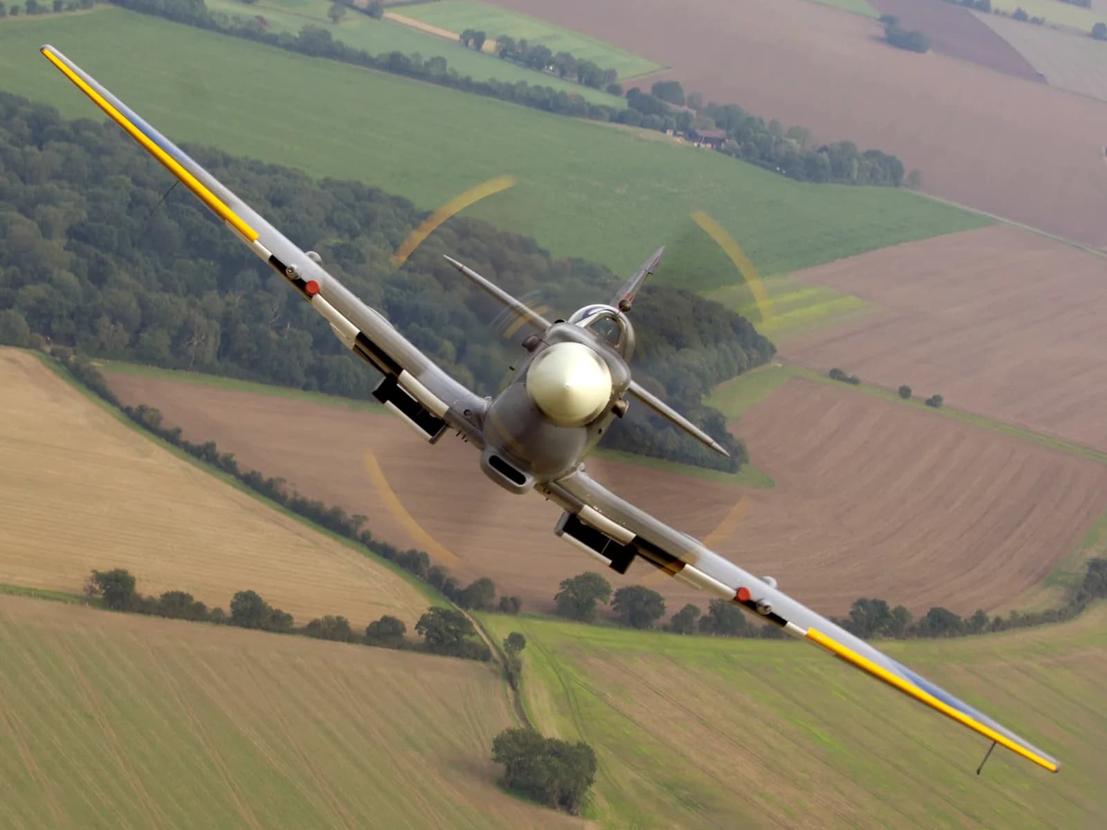 A Supermarine Spitfire airplane with a spinning propeller flies above a patchwork of green and brown agricultural fields.
