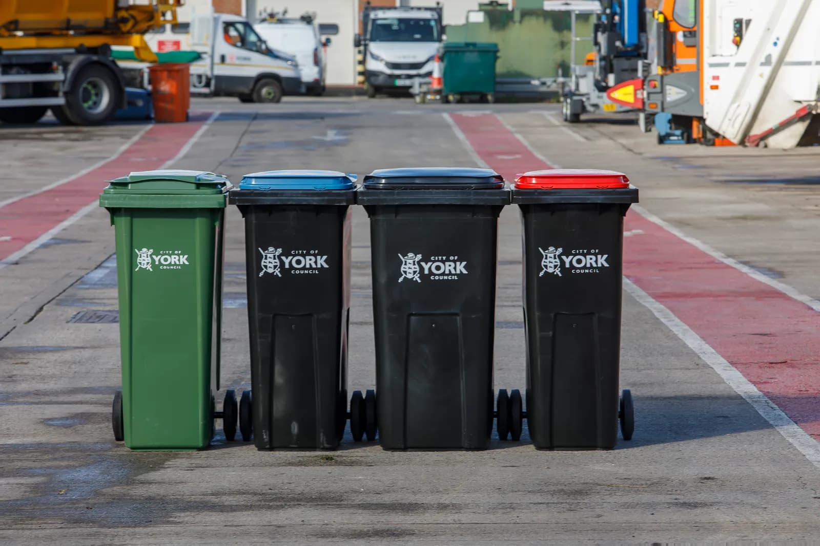 Four City of York Council recycling bins are arranged in a row in a paved area, with a green bin on the left and three black bins with blue, black, and red lids.