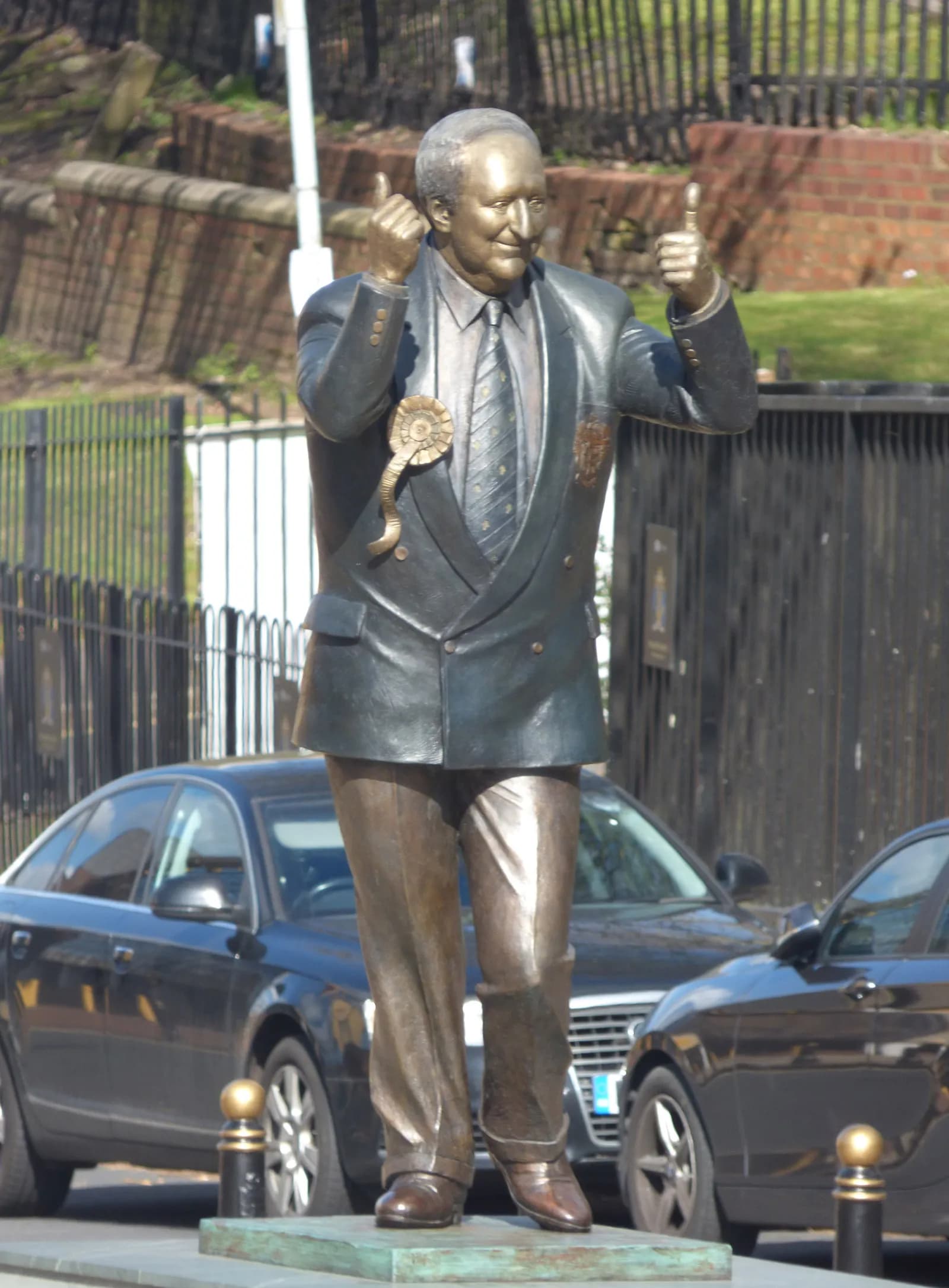 A bronze statue of a man with short hair wearing a suit, tie, and a rosette, giving two thumbs up, with cars and a fence in the blurred background.