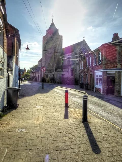 A quaint street scene features a prominent church with a tall steeple, surrounded by brick buildings and a paved sidewalk, all under a bright sky with sun rays shining through.