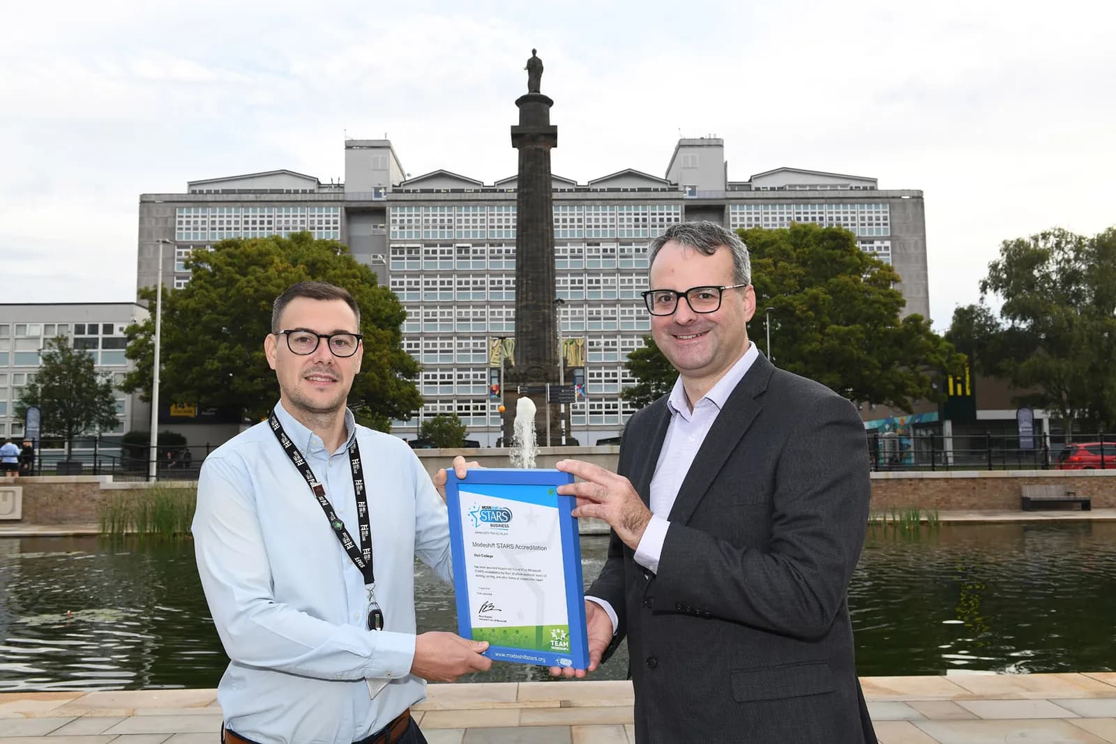 Two men in glasses hold up a blue framed certificate in front of a pond, a tall monument, and a large building.