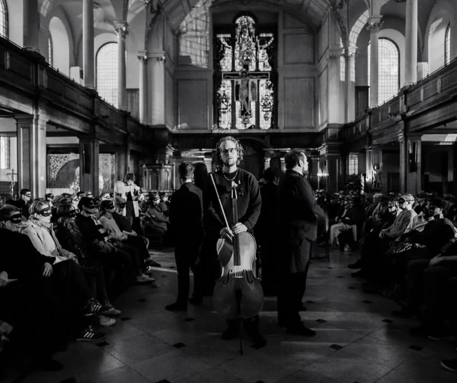 A man holding a cello stands in the middle of a church aisle, facing the viewer, flanked by seated and masked individuals on both sides.