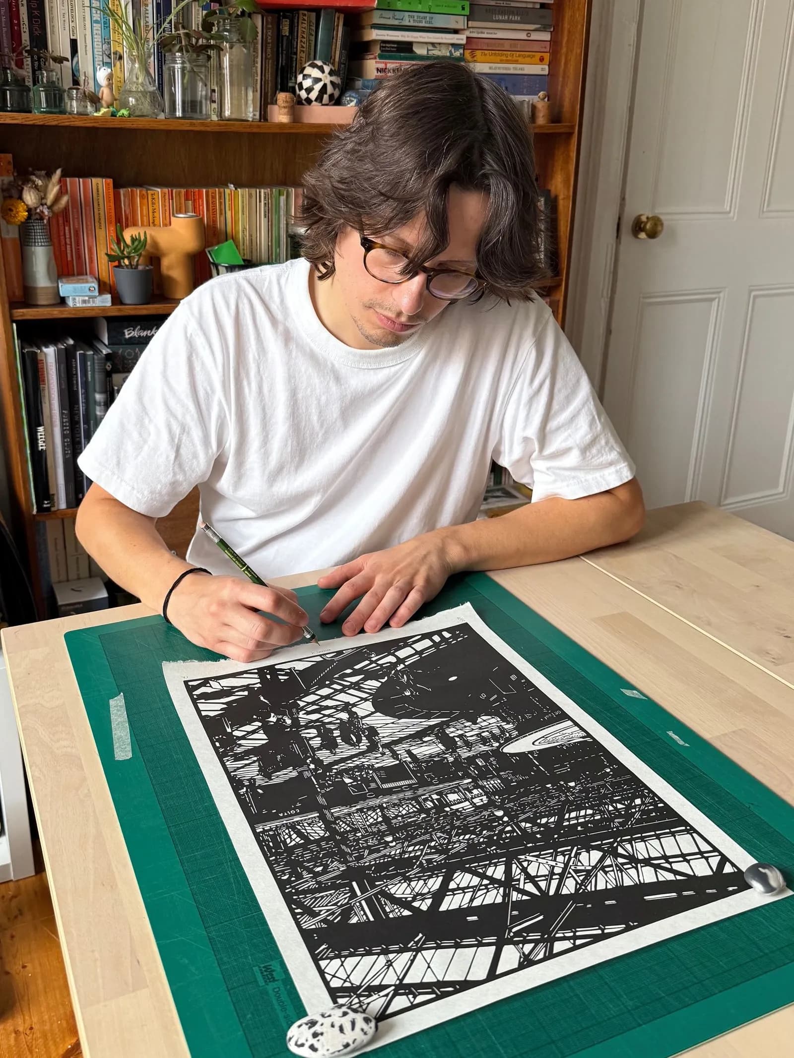 A person with long brown hair and glasses leans over a wooden table, drawing intricate black and white patterns on a large piece of white paper resting on a green mat.