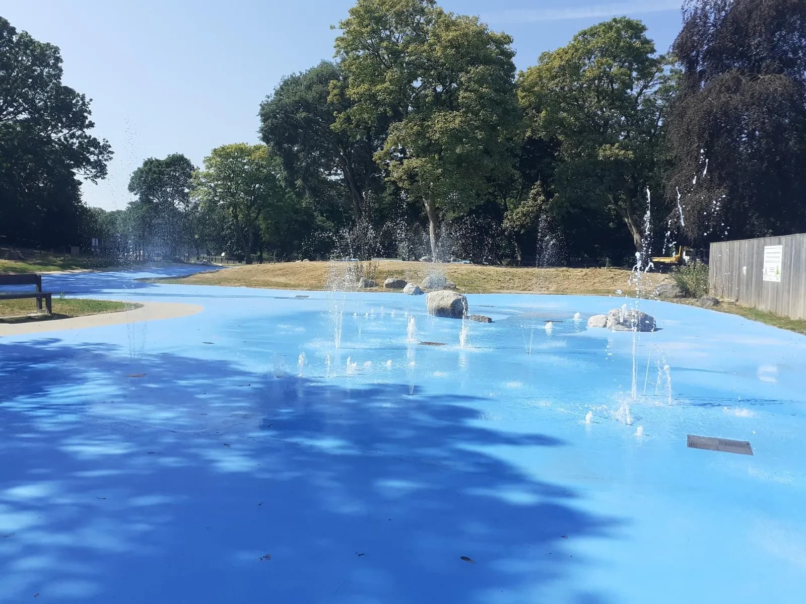A water play area with a bright blue paved surface and multiple small fountains spraying water upwards, surrounded by grassy areas and trees under a clear sky.