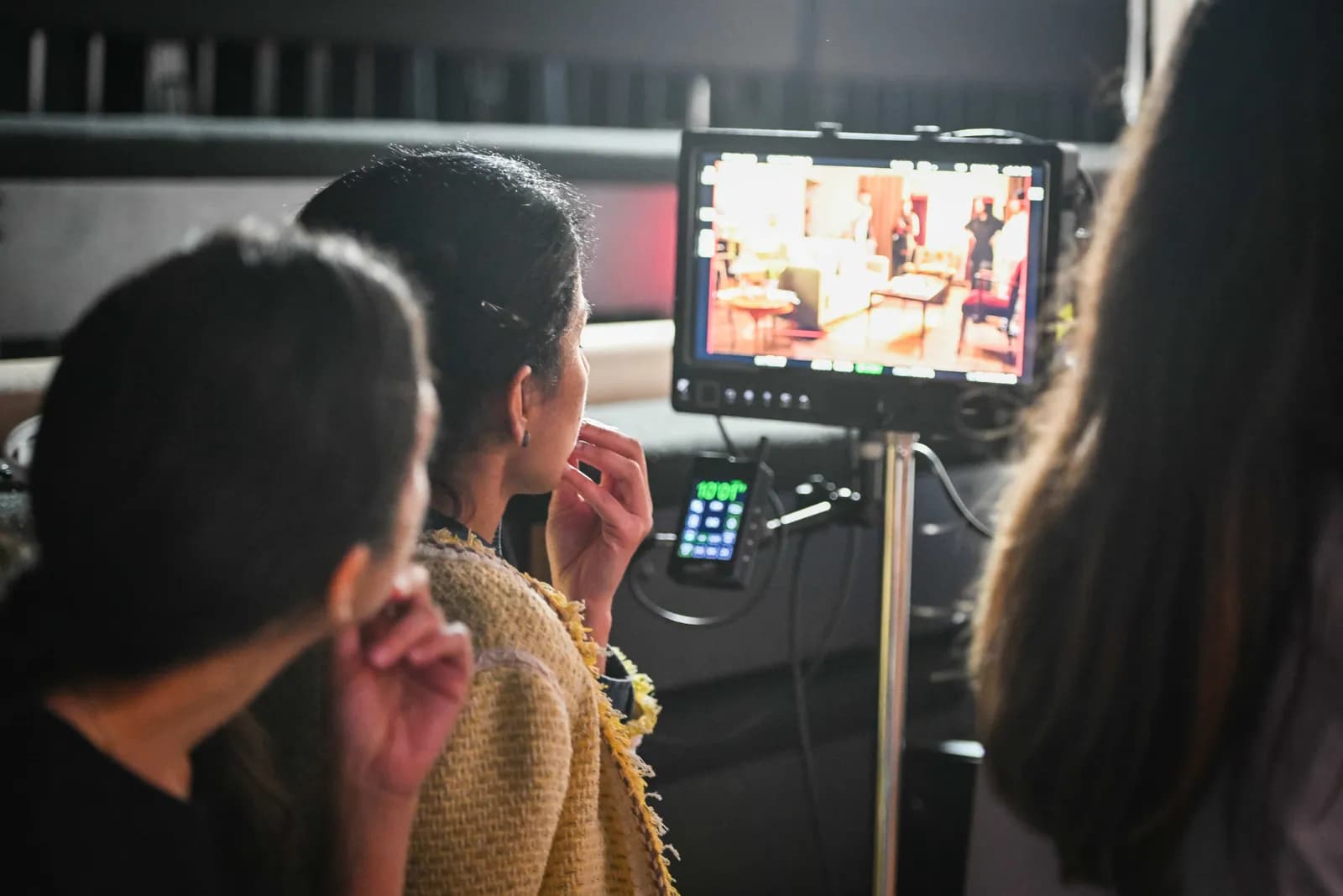 A woman from behind, with dark hair and a gold tweed jacket, intently watches a video monitor displaying an interior room scene.