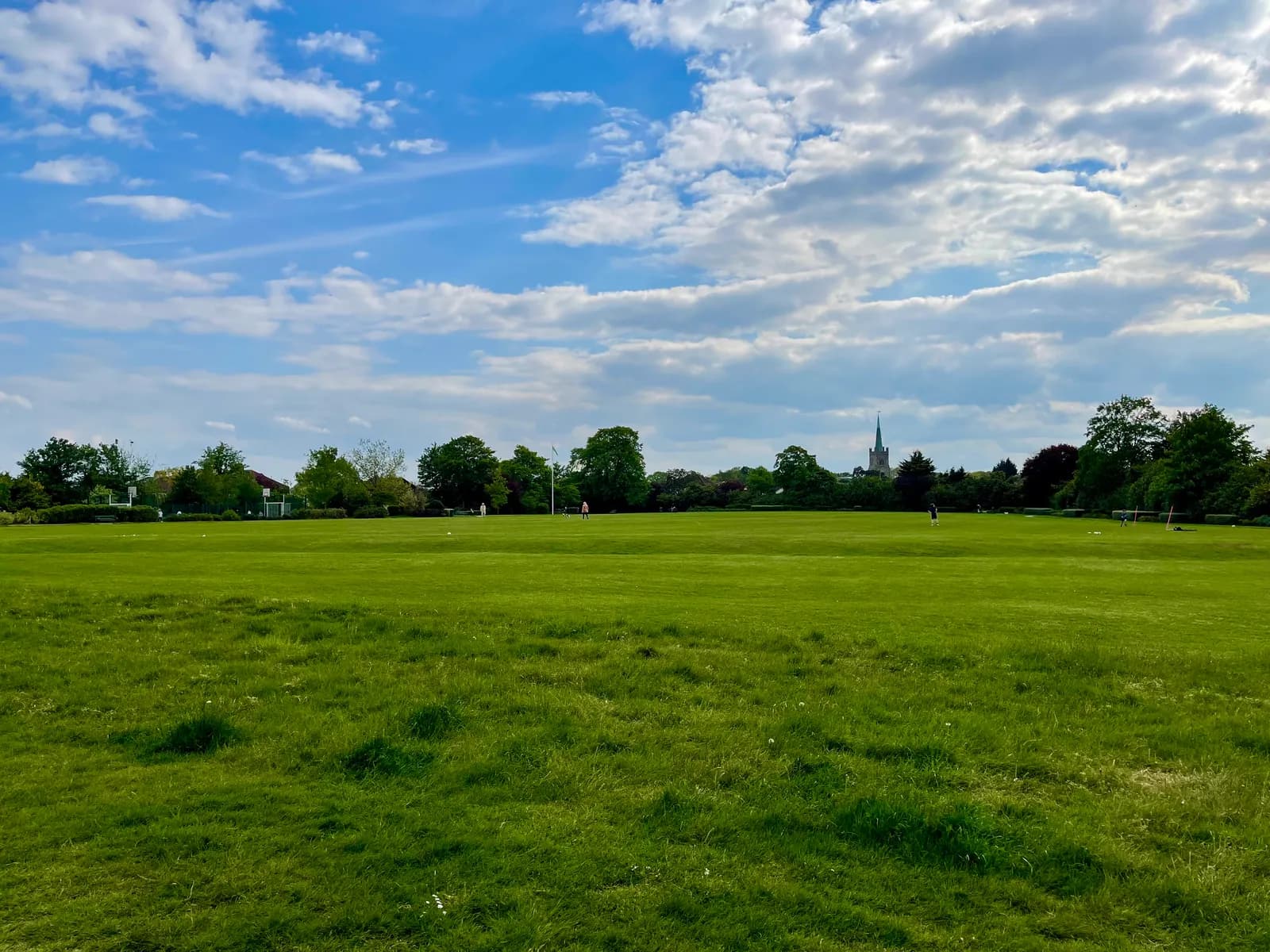 A wide, green grassy park under a partly cloudy blue sky, with trees and a distant church spire visible along the horizon.