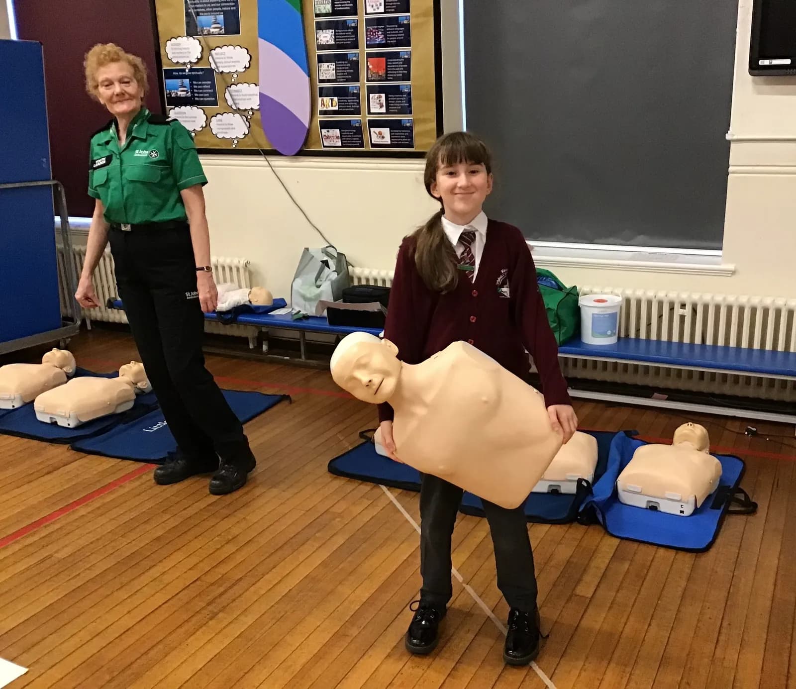 A woman in a green uniform stands next to a smiling schoolgirl holding a CPR manikin in a room with a wooden floor.