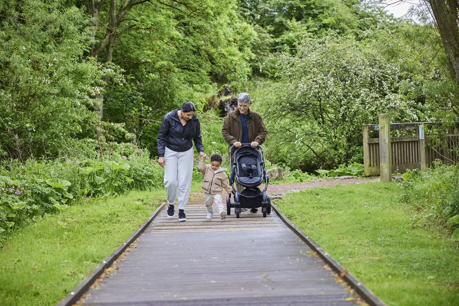 A family, including a woman holding a young child's hand and a man pushing a stroller, walks along a wooden boardwalk surrounded by lush green trees and foliage.
