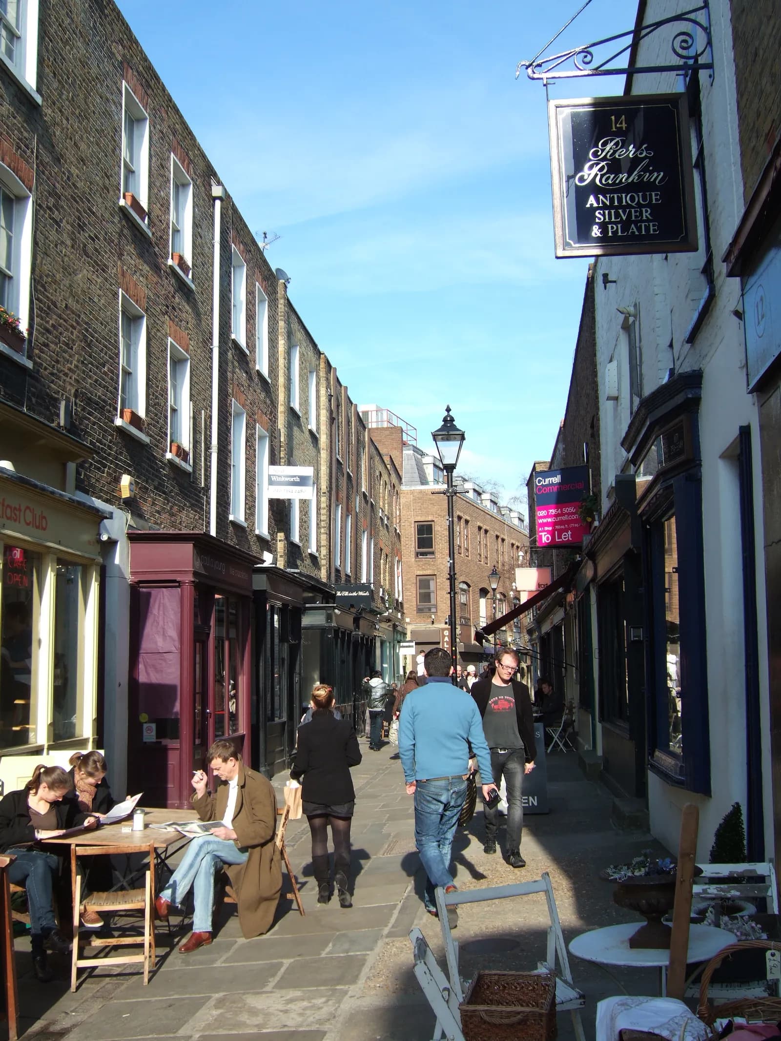 A narrow street lined with brick buildings for shops and apartments, with people walking and sitting at outdoor tables under a bright blue sky.