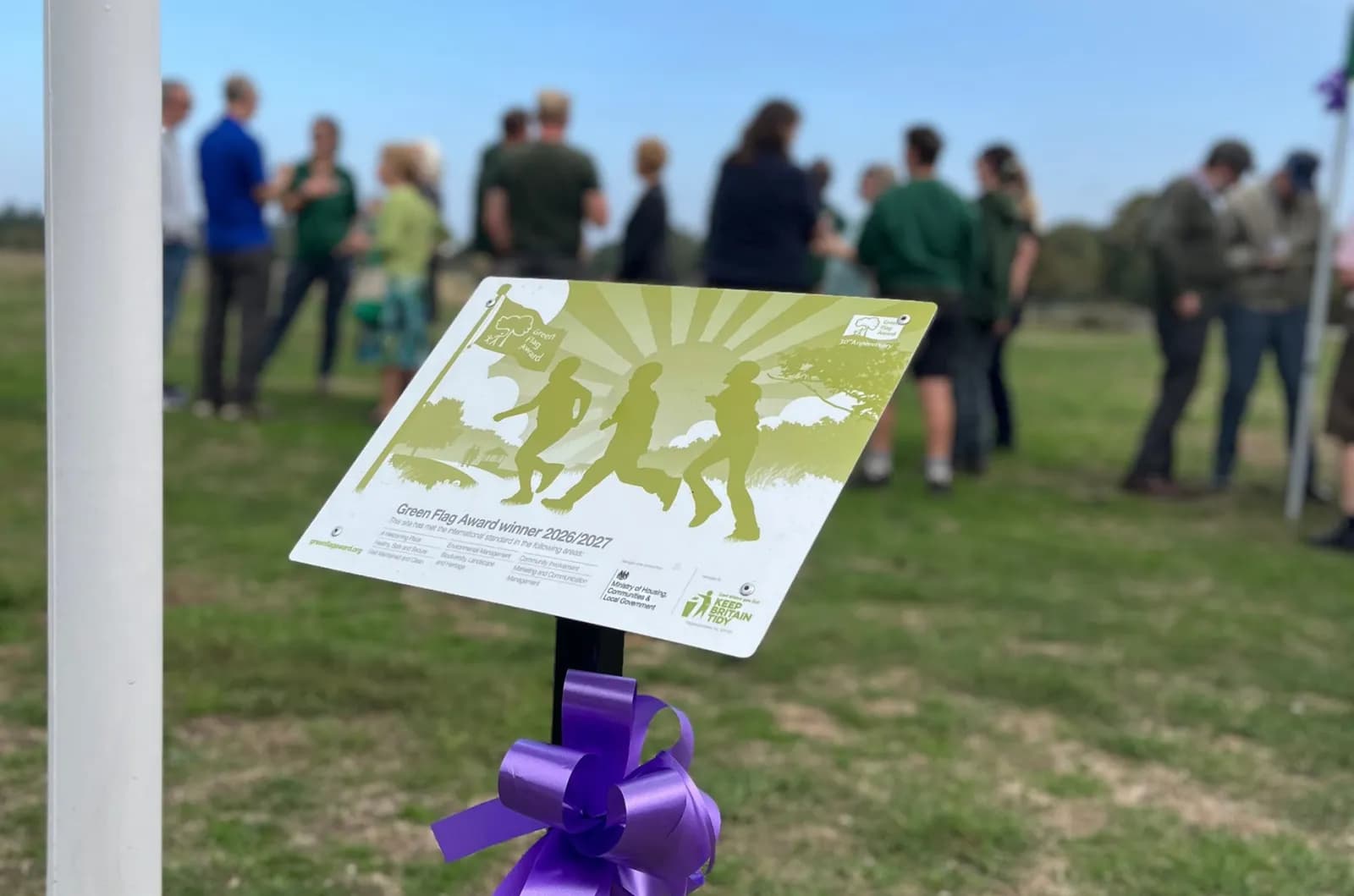A Green Flag Award winner sign for 2026/2027 with a purple bow attached, and a blurred background of people standing in a field.