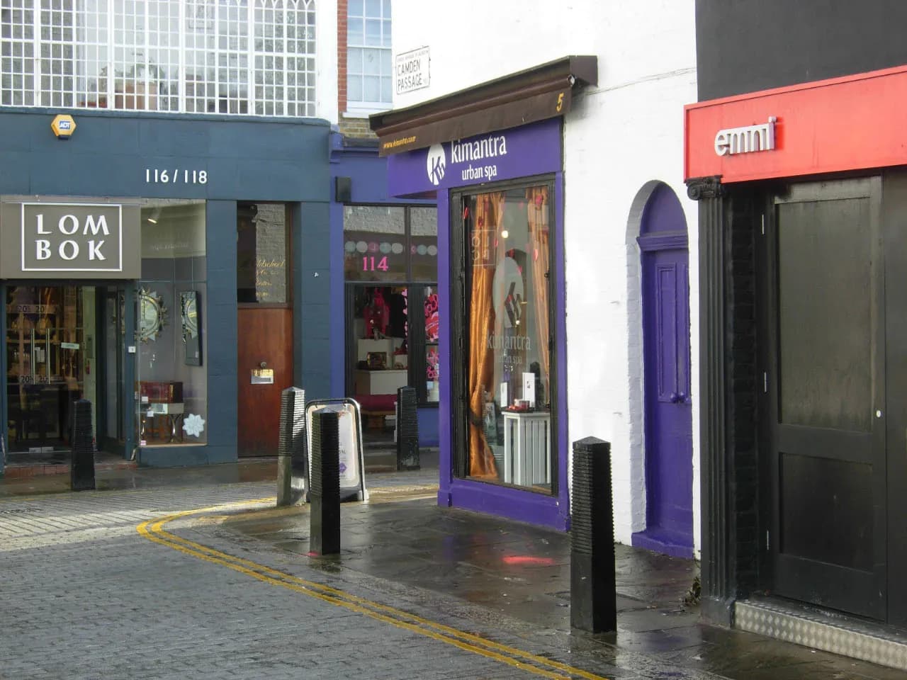 A narrow cobblestone street curves past a row of shops, including "LOMBOK," "kunantra urban spa," and "emmi."