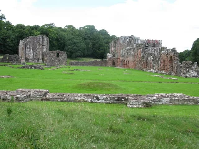 A wide-angle landscape of ancient stone ruins of an abbey set across a vast green lawn with dark green trees in the background, under a bright sky.