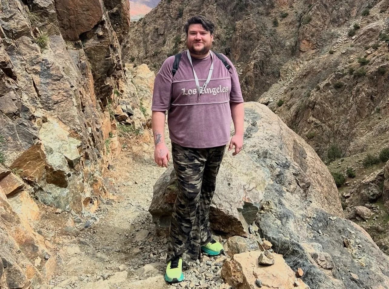 A man wearing a purple Los Angeles shirt, camouflage pants, and bright green shoes stands on a rocky mountain path.