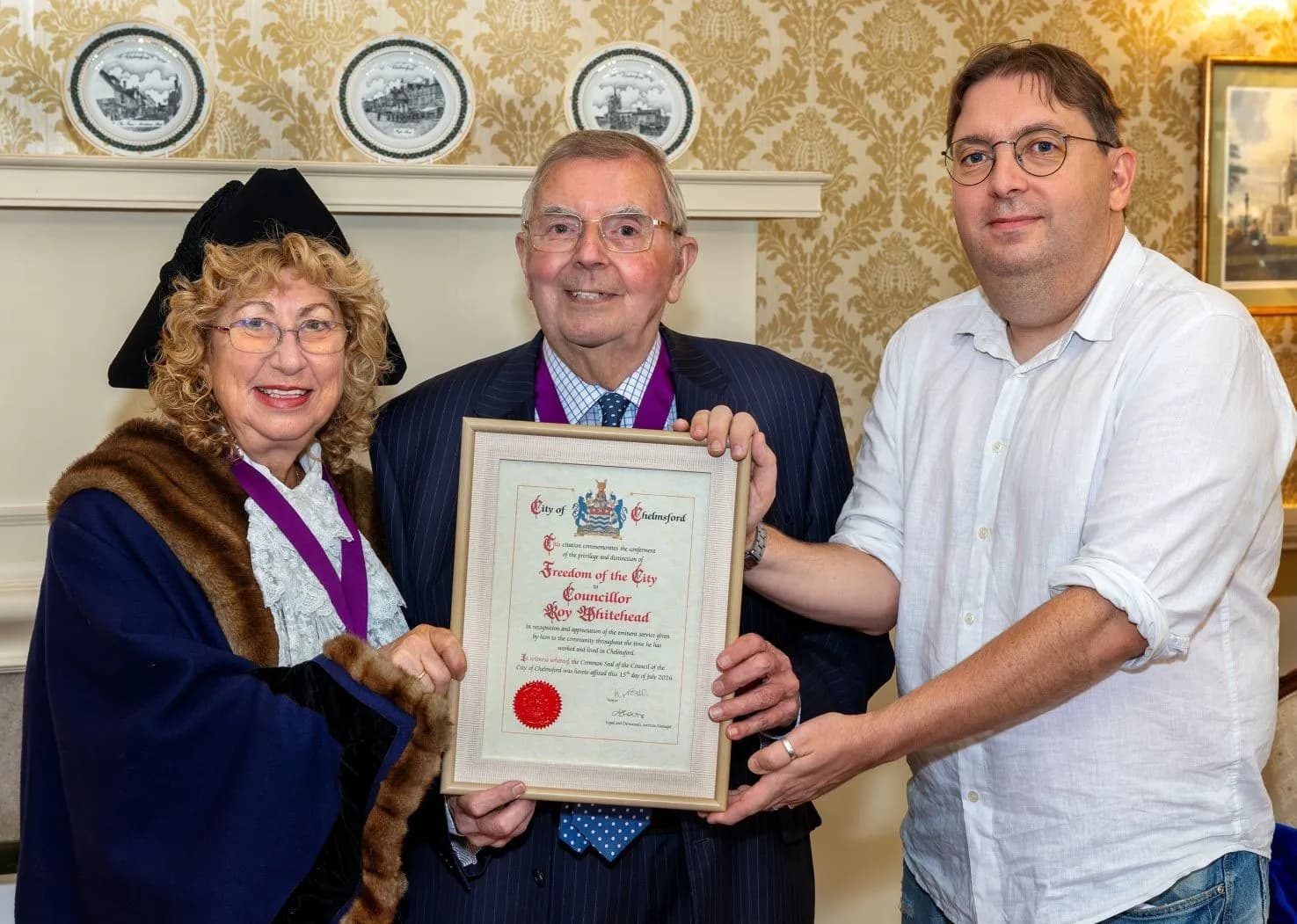 Three people stand together, two of them holding a framed certificate. The woman on the left wears a ceremonial gown and hat, the man in the middle wears a suit, and the man on the right wears a white shirt.