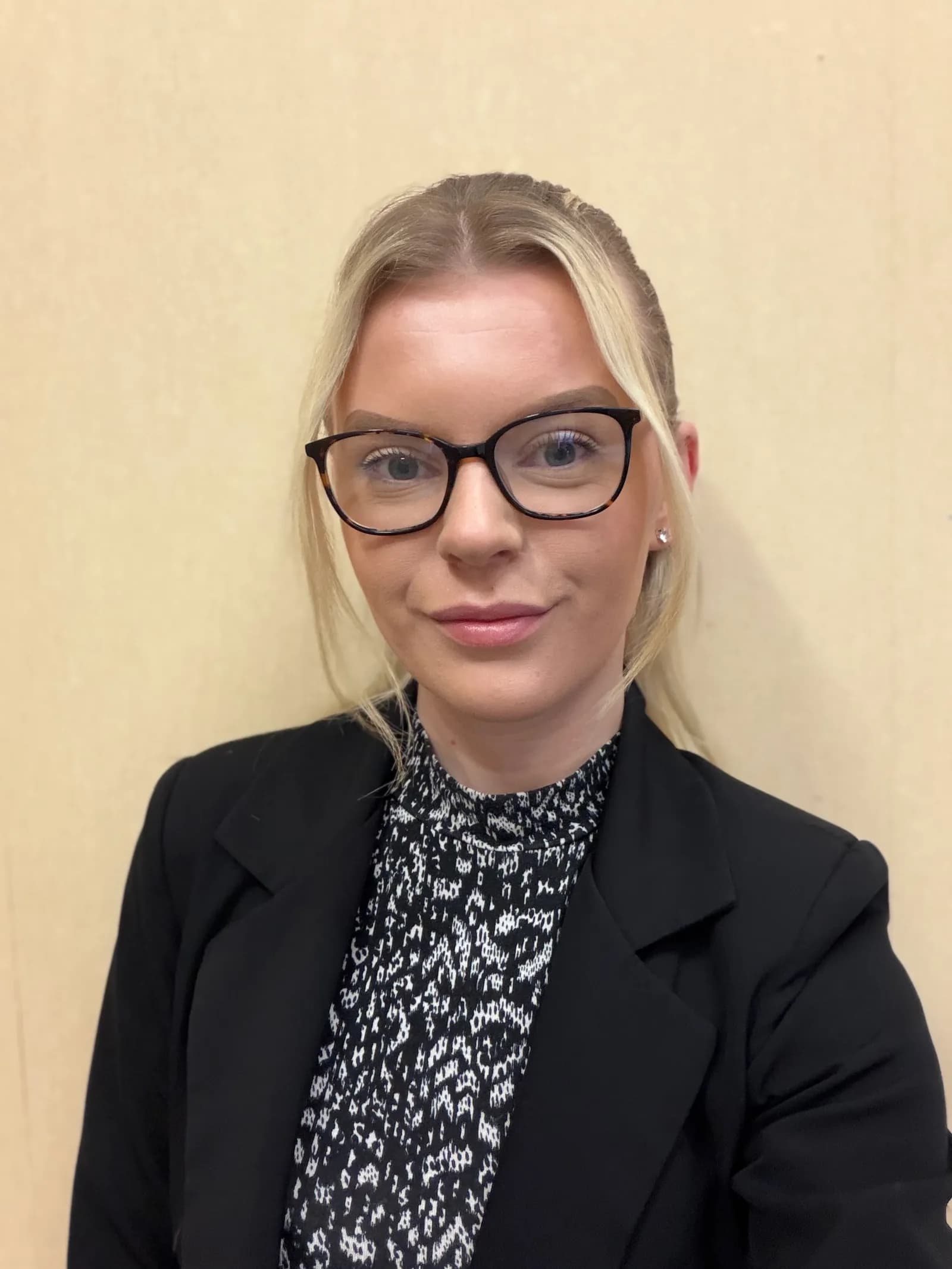 A young woman with blonde hair pulled back, wearing glasses, a black blazer, and a patterned shirt, looks directly at the viewer.