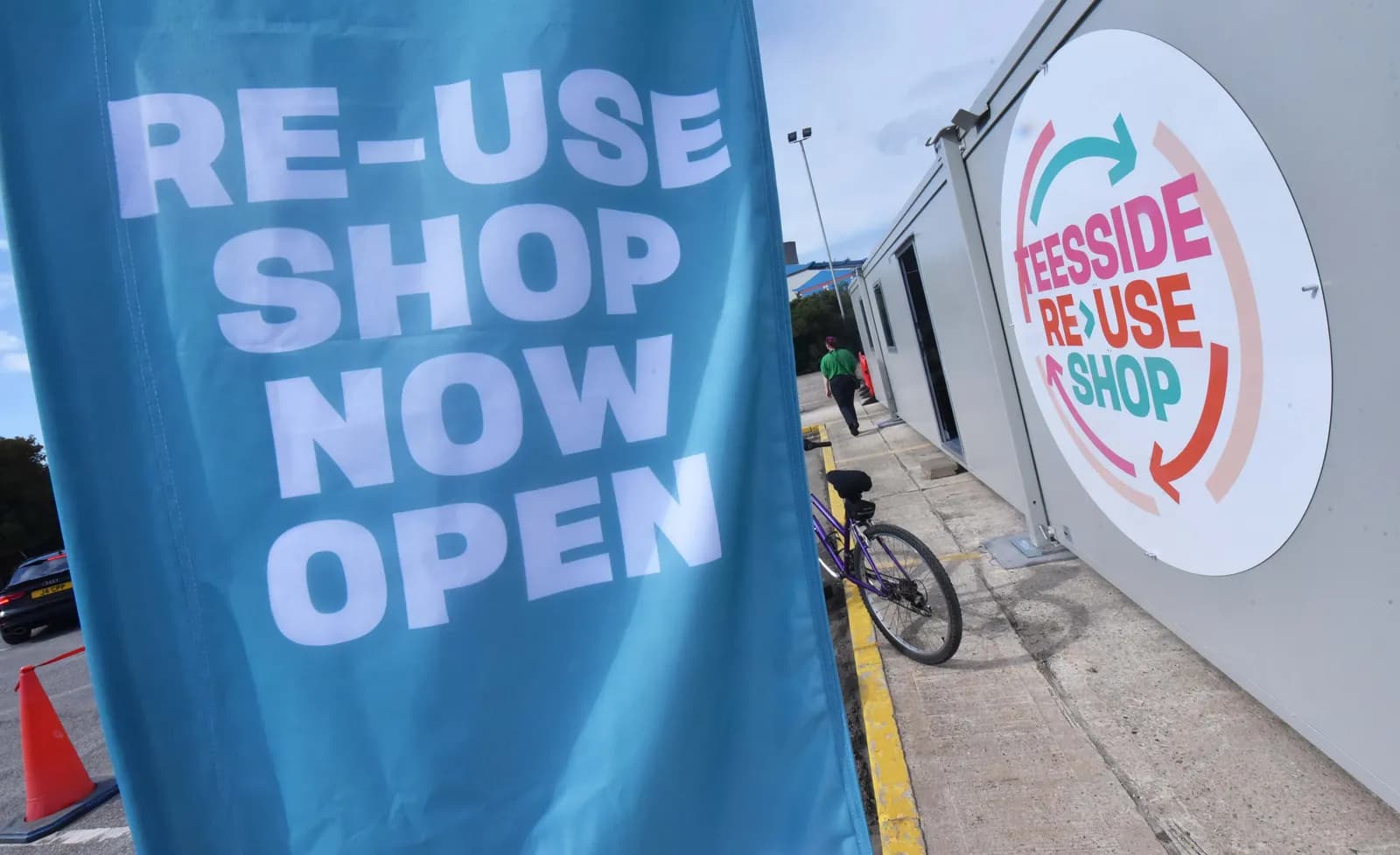 A turquoise banner with white text reads 'RE-USE SHOP NOW OPEN' in front of a white building with a round 'Teesside Re>Use Shop' logo.