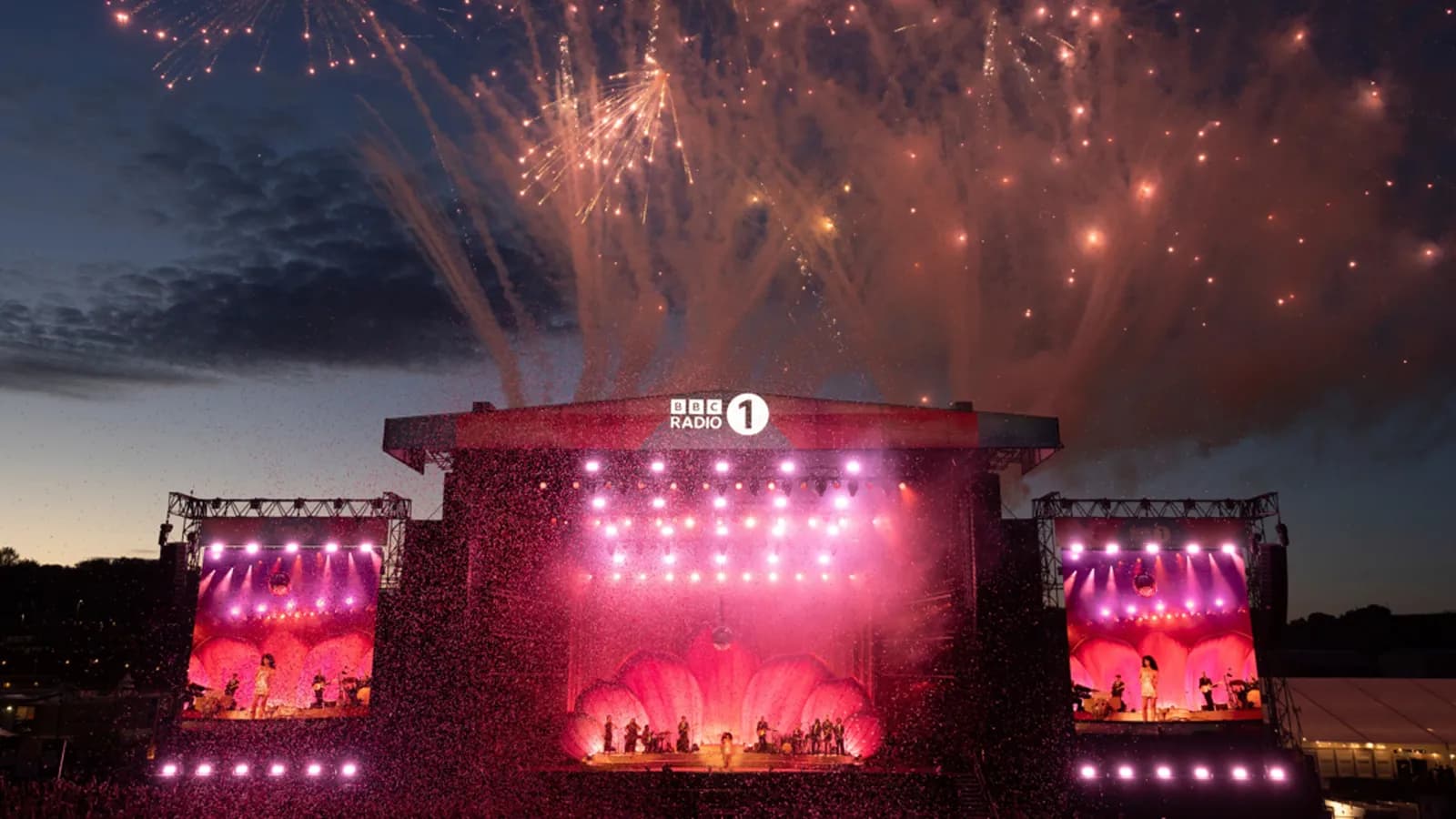 A large outdoor music stage illuminated with pink lights during a night concert, with fireworks exploding above and the BBC Radio 1 logo visible.