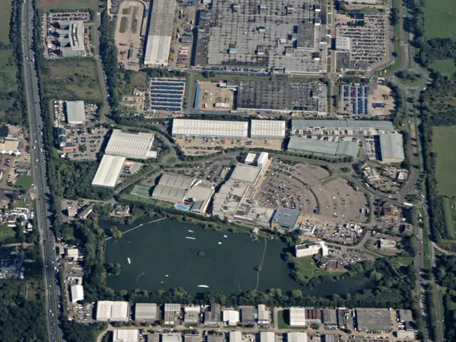 An aerial view of an industrial area with many grey warehouses, parking lots, roads, and a large dark lake surrounded by green trees on the left.