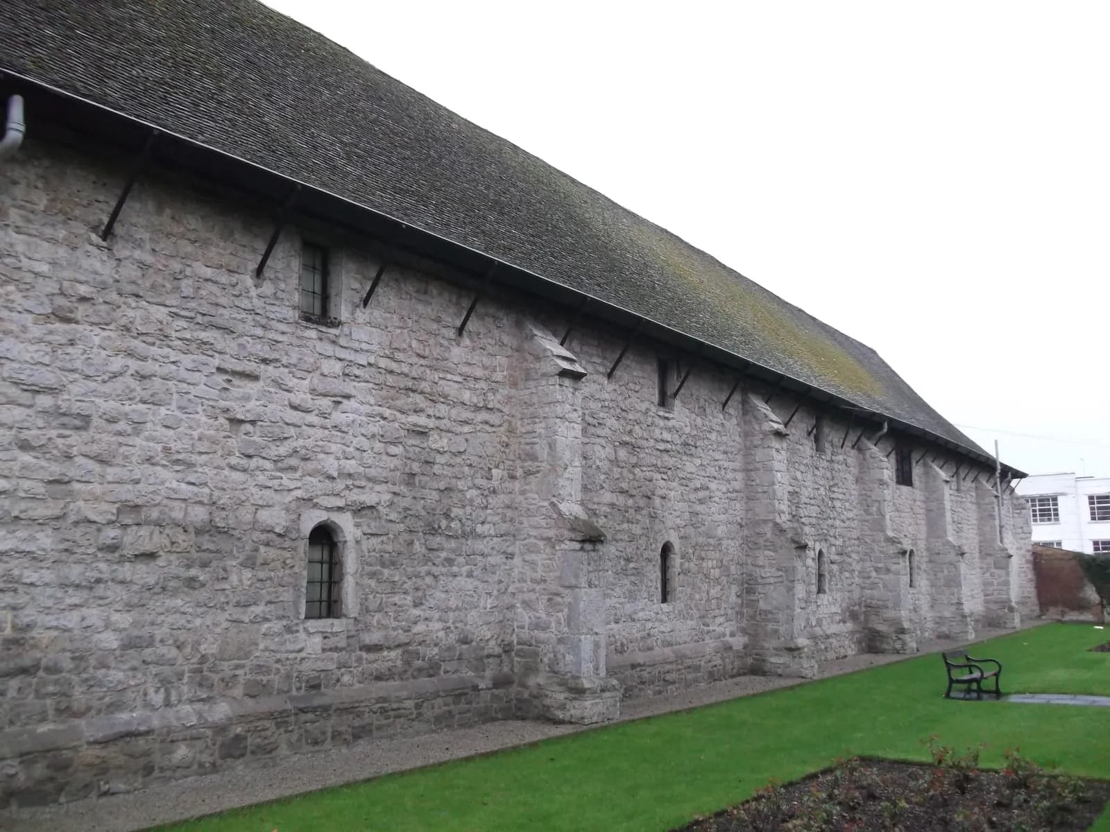 A long, weathered stone building with a dark, shingled roof, featuring several small, arched windows and buttresses, with a green lawn and a black park bench in the foreground.