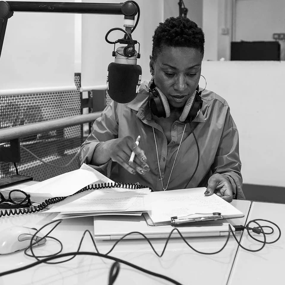 A Black woman with short hair and large hoop earrings sits at a desk with headphones around her neck, holding a pen and looking down at papers in a radio studio.