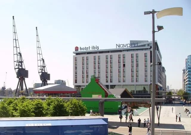 A modern cityscape featuring a hotel Ibis and a Novotel building with active cranes and greenery in the foreground, with people walking around the paved areas.