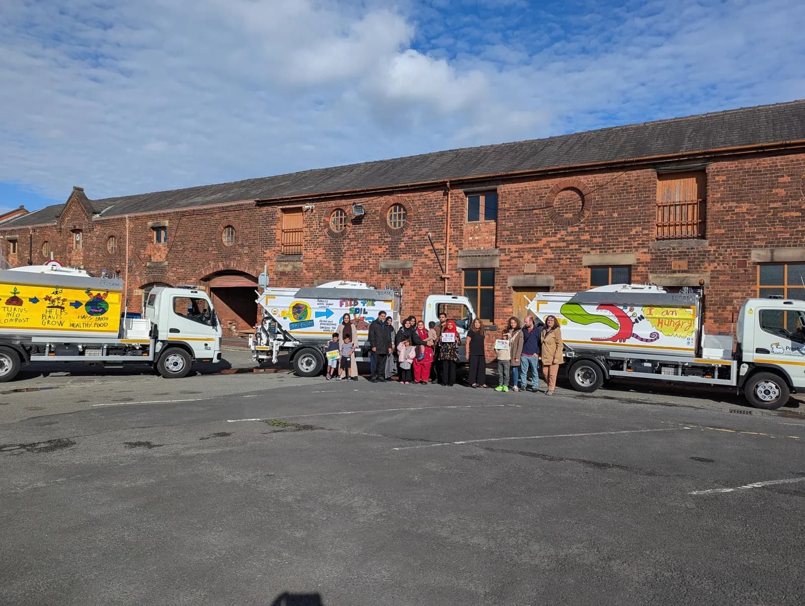 A group of people, including adults and children, stand in front of four white trucks with large tank-like backs decorated with colorful messages about food and sustainability, parked in front of a brick building.