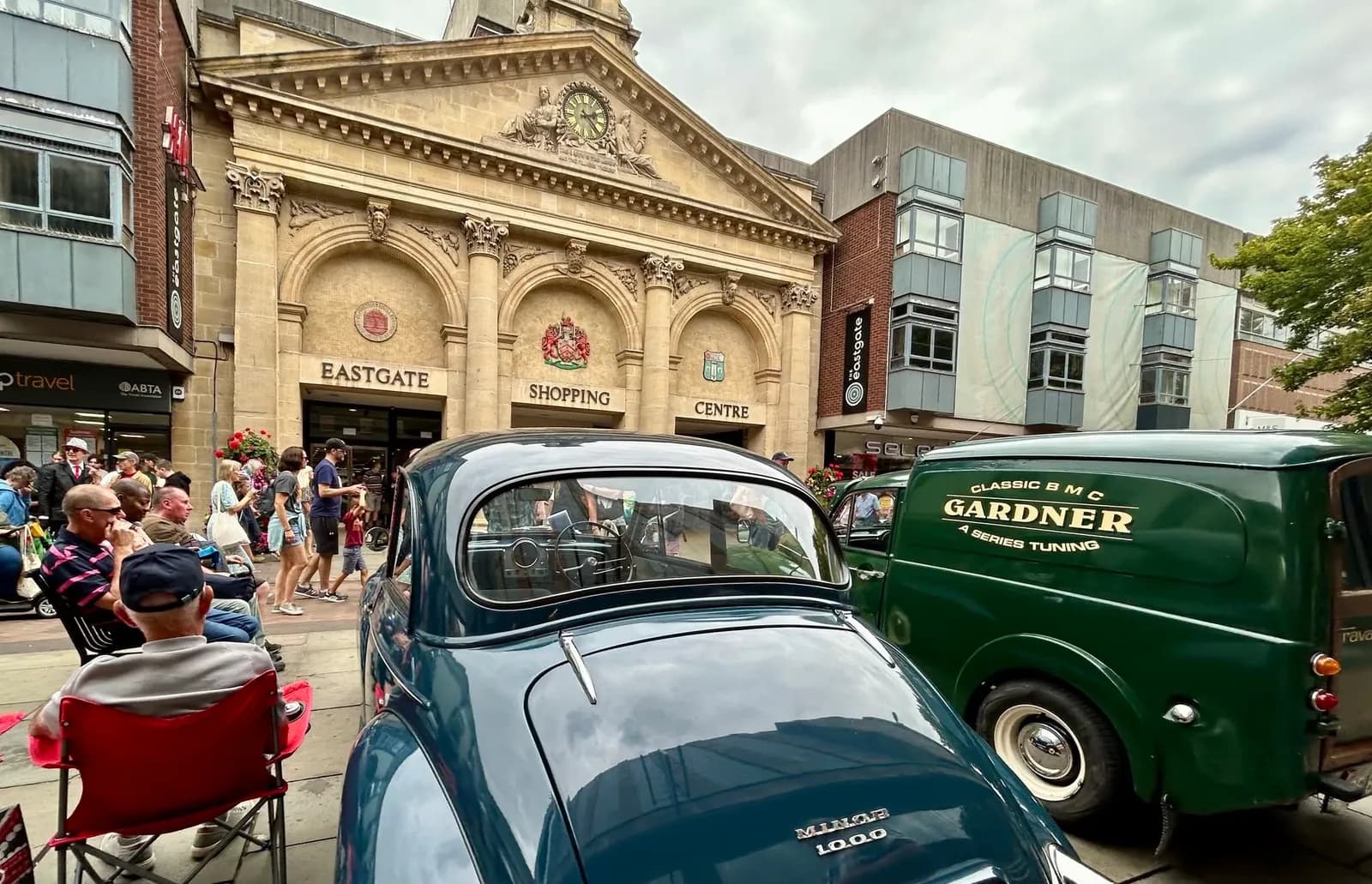 A classic blue car and green van are parked on a street in front of a building with the words "EASTGATE SHOPPING CENTRE" above its arched entrances, with people walking by.