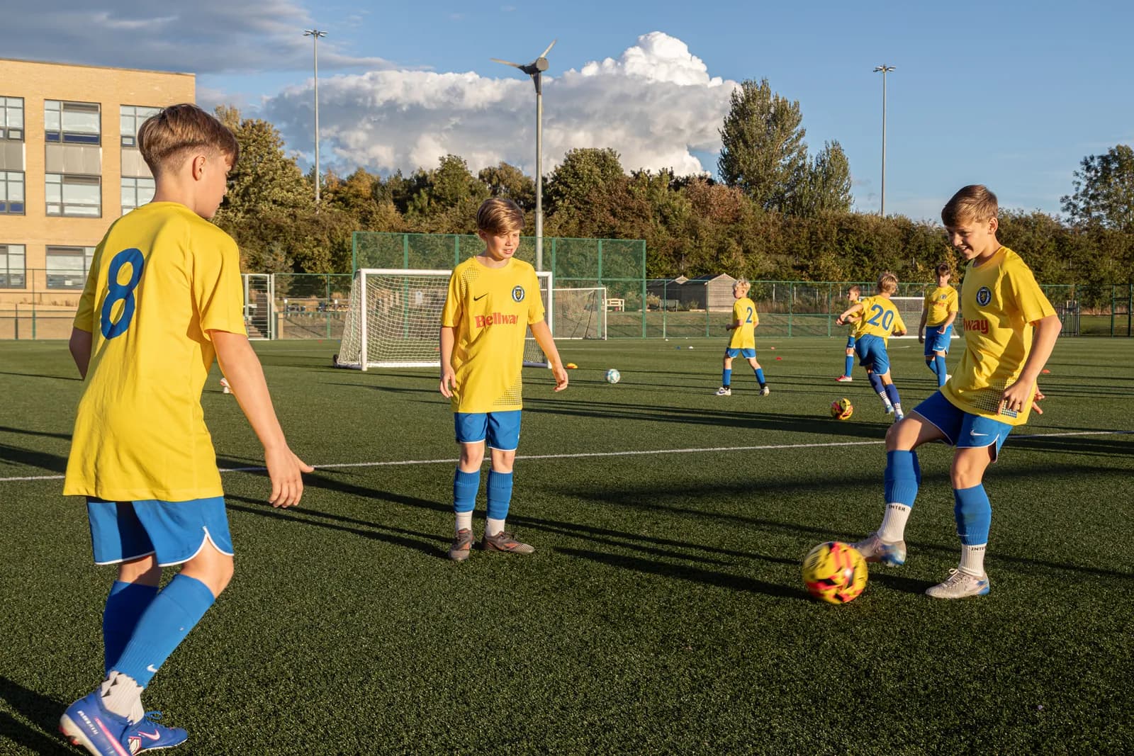 A group of boys in yellow and blue soccer uniforms playing on a green artificial turf field with a building and trees in the background.