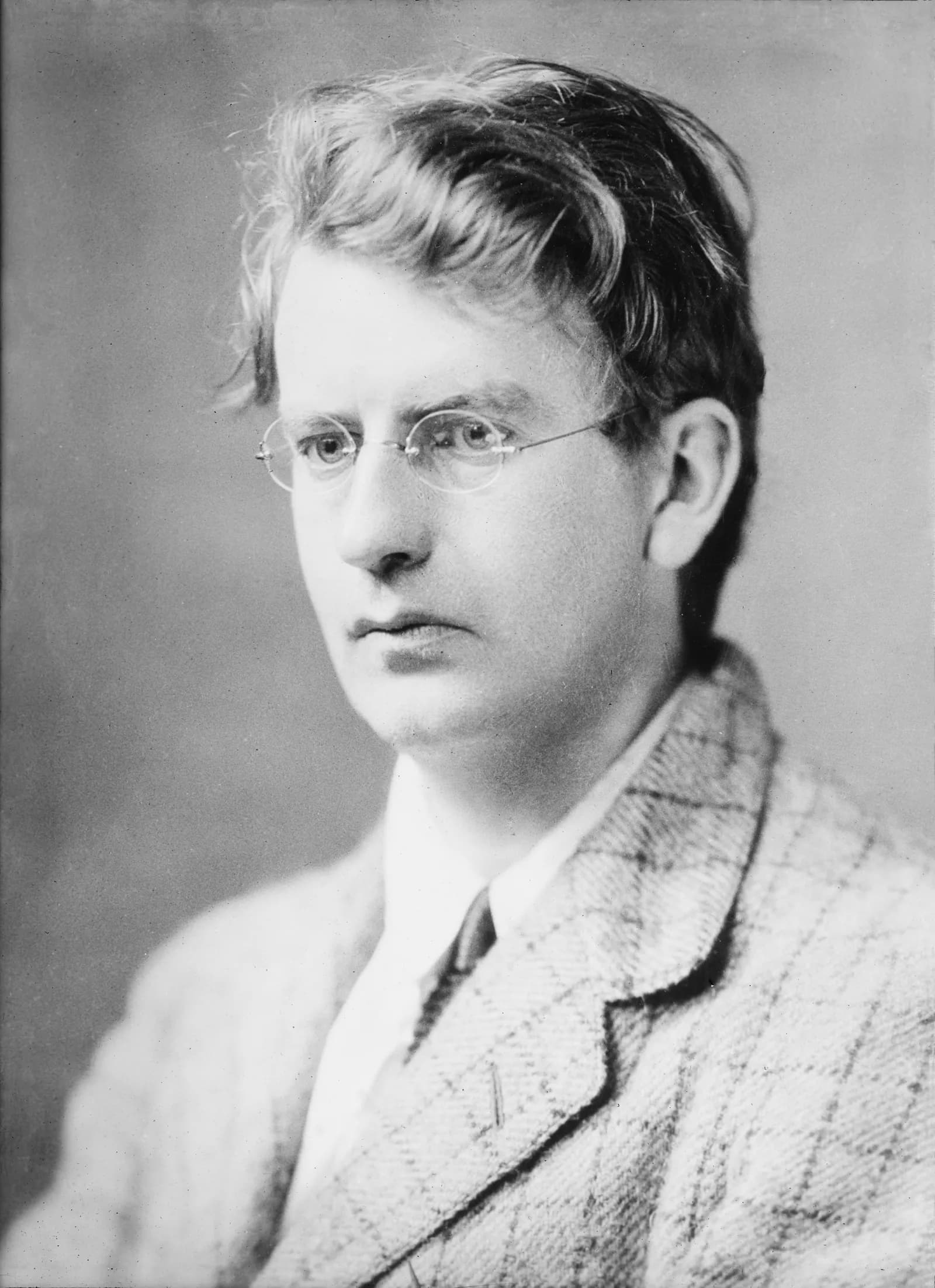 A black and white portrait of a man with tousled hair, wearing rimmed glasses and a checkered jacket, looking off to the left.