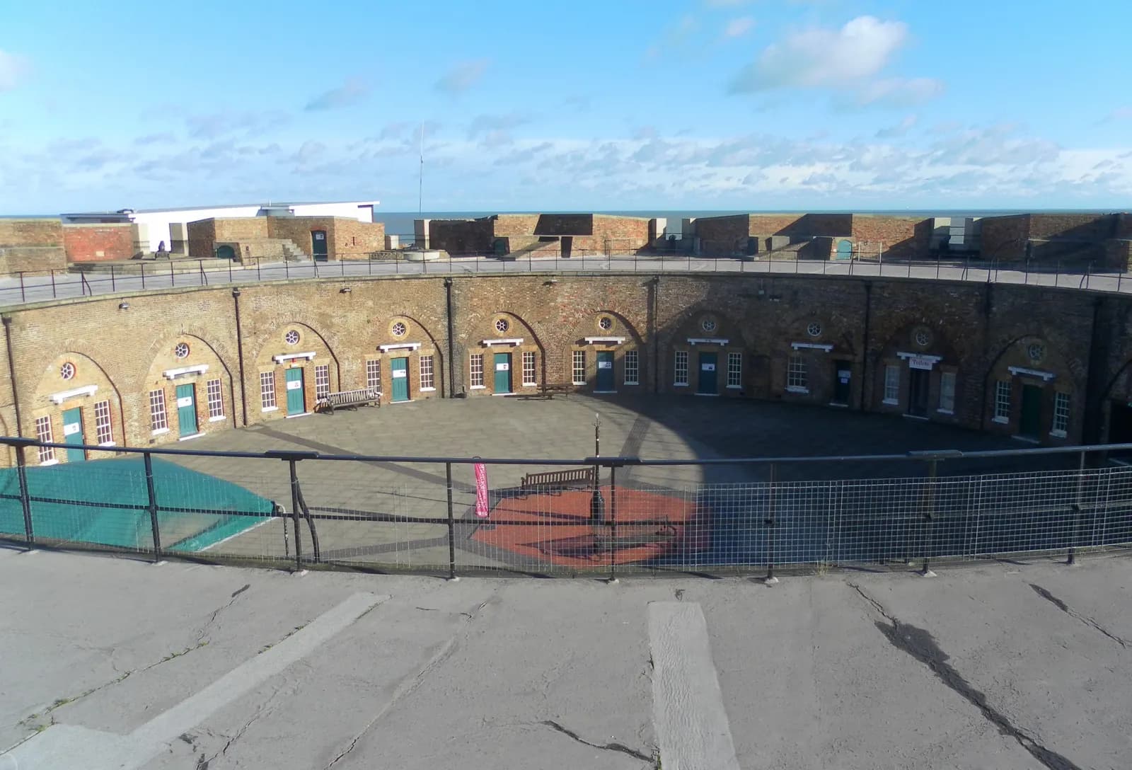 A circular, brick fort courtyard with multiple arched doorways and a concrete floor, surrounded by a low fence, under a blue sky.