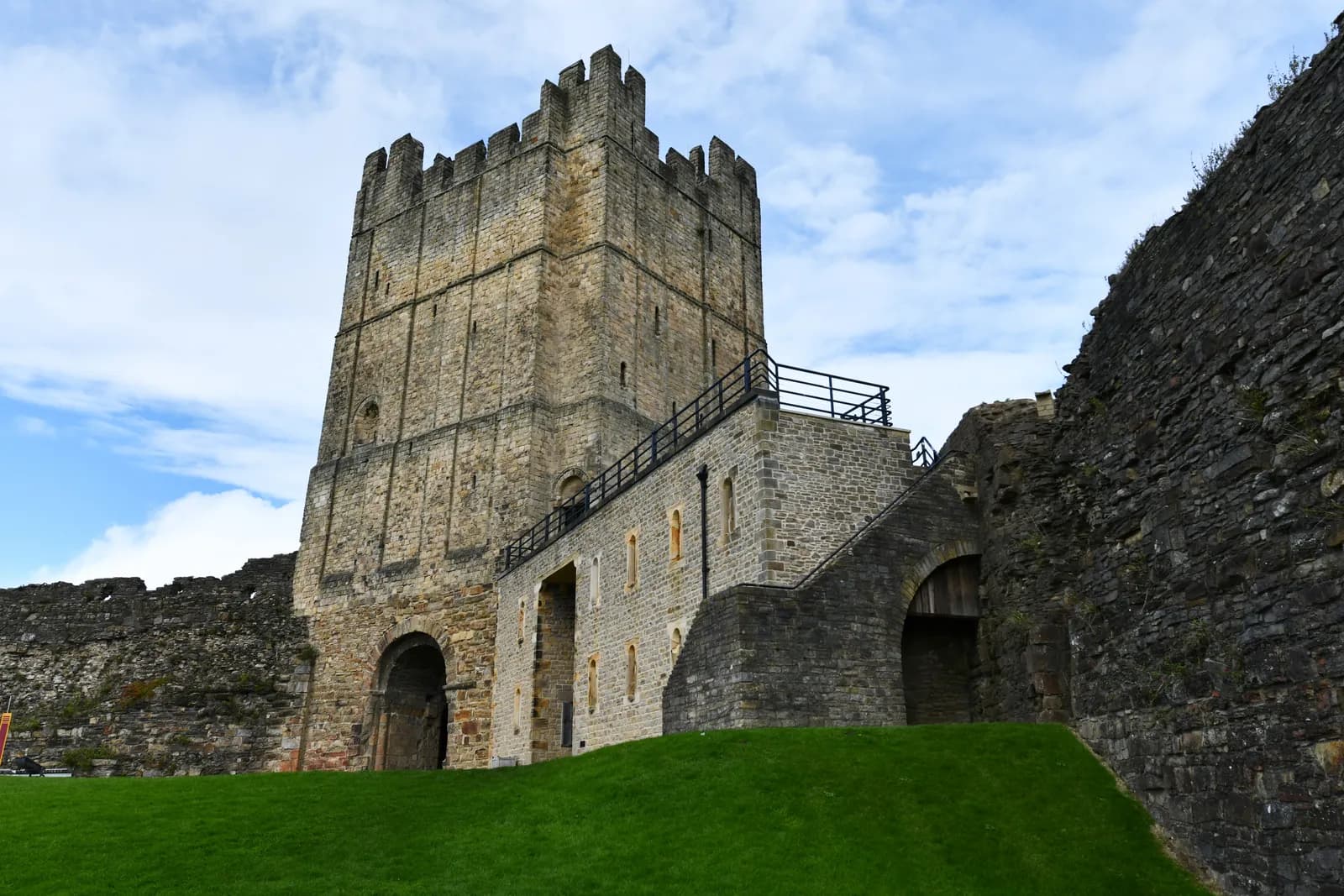 A tall, rectangular stone castle tower with battlements stands prominently against a blue sky with white clouds, with green grass sloping up to its lower walls and surrounding stone structures.