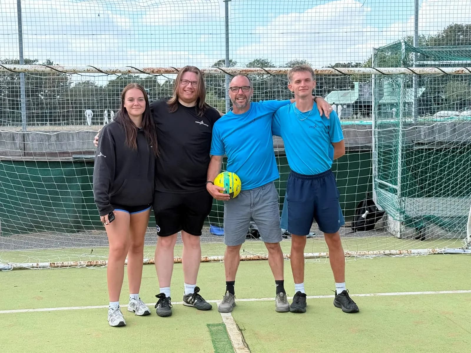A group of four people, two men and two teenagers, stand on a green sports court in front of a metal fence, smiling at the camera, with one man holding a yellow and green ball.